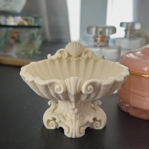 May include: Ivory-coloured, ornate seashell-shaped dish. The dish features a scalloped rim and a detailed, scrolled base. The design is reminiscent of vintage decor, perfect for holding small items. The dish is made of a solid material.
