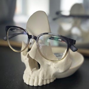 May include: Eyeglasses with black and gold frames sit on a white skull-shaped ornament. The glasses have clear lenses and the brand name "JINS" is visible. The holder is on a dark surface. A decorative item.