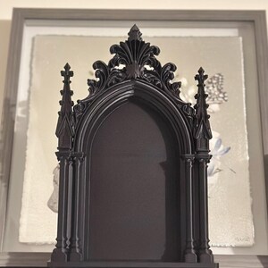 May include: A black gothic-style decorative frame with an arched opening. The frame has ornate detailing, including spires and floral accents. It is displayed on a dark surface, with framed artwork in the background.