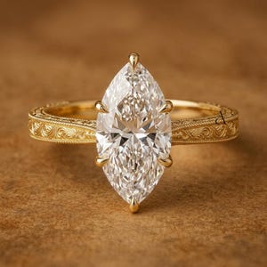 May include: A gold engagement ring with a marquise-cut diamond. The ring features a detailed, engraved band and prongs that secure the diamond. The diamond is clear and multifaceted, reflecting light. The ring is set against a warm, brown background.