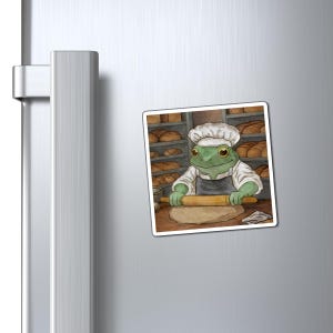 Pode incluir: Um íman quadrado com um padeiro-sapo de desenho animado, usando chapéu de chef e avental, usando um rolo em massa. Prateleiras de pão ao fundo. O sapo é verde com olhos grandes, numa cena de padaria.