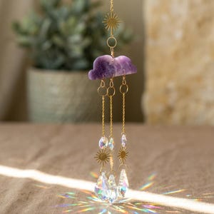 Può includere: Un acchiappasole decorativo con una nuvola di ametista viola, soli dorati e gocce di cristallo trasparente. L'acchiappasole è sospeso a una sottile catena dorata. I cristalli creano un effetto arcobaleno rifrangendo la luce.