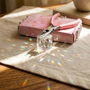 May include: A pink jewelry box with a marble design, a pair of pink-handled pliers, and a clear, faceted crystal cube. The cube is casting a rainbow of light onto the beige fabric surface.