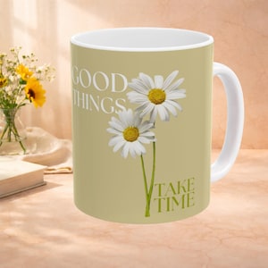 Puede incluir: Taza de cerámica blanca con fondo verde claro, con dos margaritas blancas con centros amarillos. La taza tiene las palabras "GOOD THINGS TAKE TIME" en letras blancas. Un asa blanca es visible en el lado derecho.