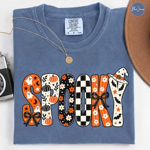 Puede incluir: Una camiseta azul de cuello redondo con la palabra SPOOKY en letras grandes y estampadas. Las letras incluyen diseños de fantasmas, calabazas, flores, cuadros blancos y negros y murciélagos. Un sombrero de bruja corona la Y y lazos negros decoran la S y la K.
