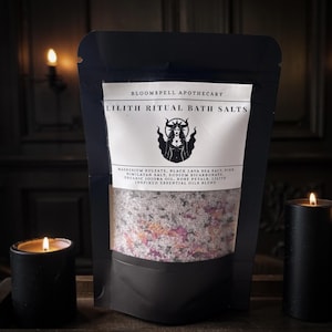 May include: Black pouch of Lilith Ritual Bath Salts by Bloomspell Apothecary. The label features a stylized illustration of a woman. The pouch contains a blend of salts and rose petals. Two black candles flank the pouch.
