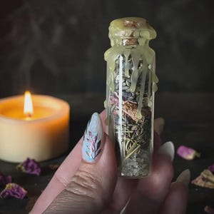 May include: A small clear glass vial filled with dried herbs, flower petals, and salt, sealed with a cork stopper covered in dripping pale yellow wax. The contents are layered with green, pink, and white textures visible through the glass.
