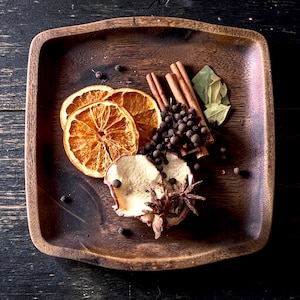 May include: A square wooden tray containing a collection of dried spices and fruits, including two orange slices, three cinnamon sticks, a cluster of whole black peppercorns, two star anise, a dried apple slice, and several dried bay leaves.