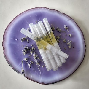 Op de afbeelding: Zes transparante nageloliepennen gevuld met gele vloeistof en kruiden, gerangschikt op een paarse en witte agaat schijf. Gedroogde lavendel takjes zijn verspreid rond de pennen. De pennen hebben witte doppen en applicators.