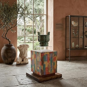 May include: An interior shot of a room with a stone floor and a large window. A colorful, wooden block stand holds a bronze vessel. A white sculpture and a potted olive tree are also visible. A glass display case is in the background.