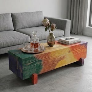 Könnte beinhalten: Ein rechteckiger Couchtisch aus einem massiven Holzbalken mit einem lebendigen Farbverlauf, der von Dunkellila und Grün an einem Ende zu hellem Gelb und Orange am anderen Ende übergeht und auf vier kleinen Blockfüßen steht.