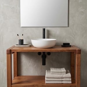 May include: A wooden bathroom vanity featuring a white vessel sink, a tall black faucet, and a black drain pipe. The vanity has a lower shelf holding a stack of folded beige towels. A dark gray toothbrush holder and a black soap bar are on top.