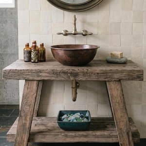 Puede incluir: Un mueble de baño de madera rústica con un lavabo de cobre martillado, un grifo de bronce montado en la pared y un estante inferior con un cuenco cuadrado de color verde azulado lleno de vidrio marino. Tres botellas de vidrio ámbar y jabón.