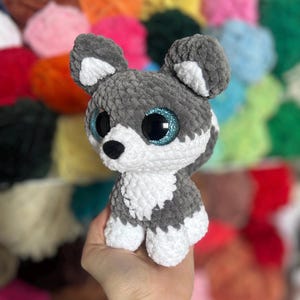 May include: A handmade plush husky dog with gray and white fur, large blue glitter eyes, and a black nose. The stuffed animal is crocheted and has a soft, textured appearance. The background is a colorful array of yarn.