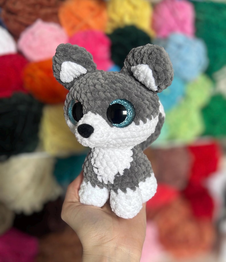 May include: A handmade plush husky dog with gray and white fur, large blue glitter eyes, and a black nose. The stuffed animal is crocheted and has a soft, textured appearance. The background is a colorful array of yarn.