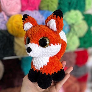 May include: A handmade orange, white, and black crocheted fox plush toy with large, glittery golden eyes. The fox has a fluffy texture and is sitting upright. The background features colorful yarn balls.