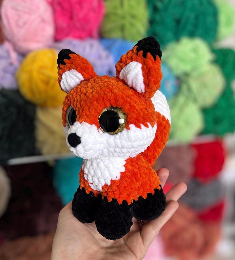 May include: A handmade orange, white, and black crocheted fox plush toy with large, glittery golden eyes. The fox has a fluffy texture and is sitting upright. The background features colorful yarn balls.