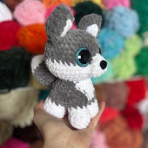 May include: A handmade crochet husky dog plushie in gray and white. The plush toy has large, sparkly blue eyes and a black nose. The dog is sitting upright, with its ears perked up. The background is blurred, showing colorful yarn.