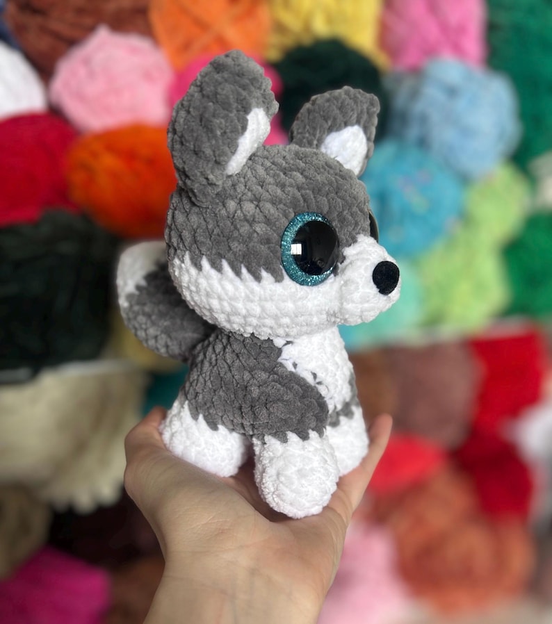 May include: A handmade crochet husky dog plushie in gray and white. The plush toy has large, sparkly blue eyes and a black nose. The dog is sitting upright, with its ears perked up. The background is blurred, showing colorful yarn.