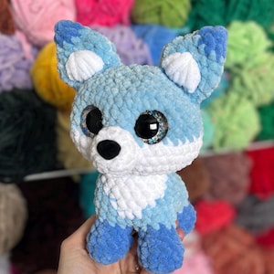 May include: A handmade blue and white crocheted fox plush toy with large, glittery eyes. The fox has a fluffy texture and is held in a person's hand. The background features a variety of colorful yarn skeins.