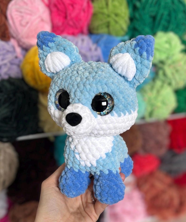 May include: A handmade blue and white crocheted fox plush toy with large, glittery eyes. The fox has a fluffy texture and is held in a person's hand. The background features a variety of colorful yarn skeins.