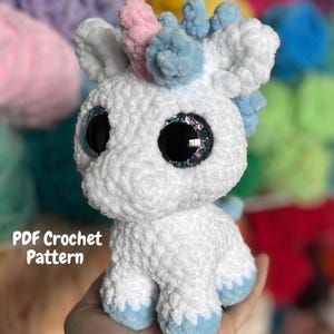 May include: A white crocheted unicorn with blue and pink accents. The plush toy has large, glittery eyes and blue hooves. The text "PDF Crochet Pattern" is visible on the image.
