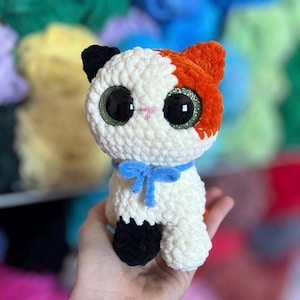 May include: A handmade crochet cat doll with white, orange, and black details. The cat has large, glittery green eyes, a blue bow, and is held in a hand. The background features colorful yarn.