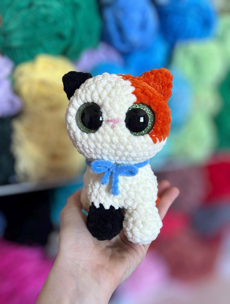 May include: A handmade crochet cat doll with white, orange, and black details. The cat has large, glittery green eyes, a blue bow, and is held in a hand. The background features colorful yarn.