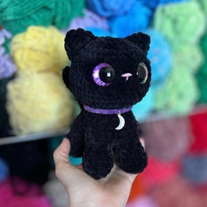 May include: A handmade black crochet cat doll with large, iridescent purple eyes. The cat wears a purple collar with a silver crescent moon charm. The background features colorful yarn skeins.