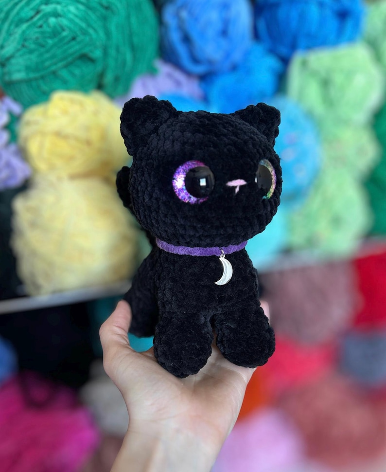 May include: A handmade black crochet cat doll with large, iridescent purple eyes. The cat wears a purple collar with a silver crescent moon charm. The background features colorful yarn skeins.