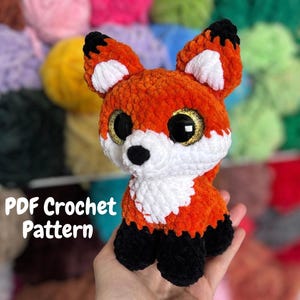 Puede incluir: Un zorro de ganchillo naranja, blanco y negro hecho a mano con grandes ojos dorados. El zorro tiene una textura esponjosa y está sentado erguido. El texto "PDF Crochet Pattern" es visible en la imagen.