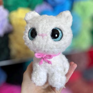 May include: A handmade white plush cat with large, glittery blue eyes and a pink bow. The cat is made of soft, fluffy material and has a sweet, innocent expression. The background is blurred, with colorful yarn in various shades.