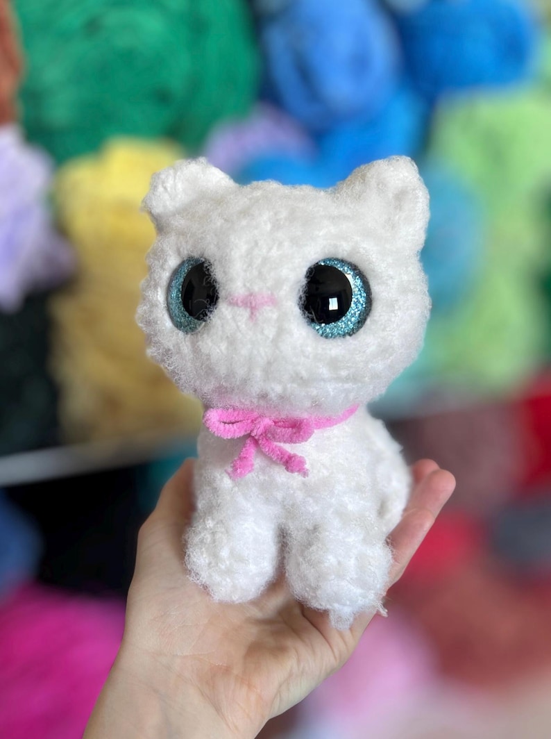 May include: A handmade white plush cat with large, glittery blue eyes and a pink bow. The cat is made of soft, fluffy material and has a sweet, innocent expression. The background is blurred, with colorful yarn in various shades.