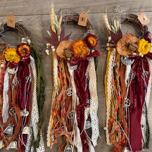 May include: Three decorative wall hangings featuring circular grapevine wreaths adorned with dried flowers, wheat, and leaves. Each wreath has long, cascading ribbons in shades of burgundy, orange, green, and cream, accented with metal charms and bells.