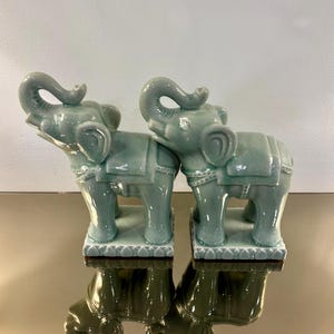 May include: Two matching celadon green ceramic elephant figurines standing on rectangular bases. Each elephant features a raised trunk, a saddle blanket with decorative trim, and a smooth, glossy glaze finish with subtle crackle detailing.