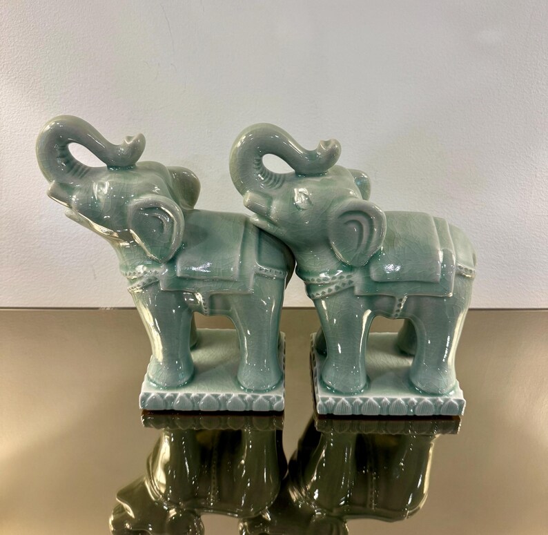 May include: Two matching celadon green ceramic elephant figurines standing on rectangular bases. Each elephant features a raised trunk, a saddle blanket with decorative trim, and a smooth, glossy glaze finish with subtle crackle detailing.