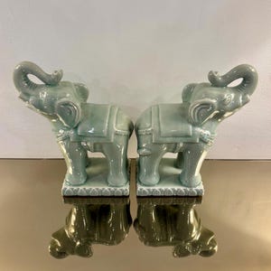 May include: A pair of celadon green ceramic elephant figurines standing on rectangular bases with lotus petal detailing. Each elephant is depicted with its trunk raised upward, wearing a saddle blanket, and featuring a glossy glazed finish.