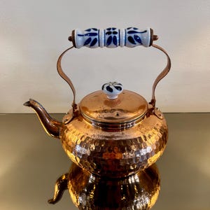 May include: Copper tea kettle with a hammered texture. The kettle features a white and blue ceramic handle and a matching lid knob. The spout and handle supports are copper-colored. A decorative kitchen item.