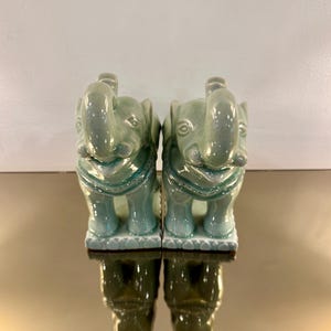 May include: A pair of matching ceramic elephant figurines featuring a glossy, light green celadon glaze. Each elephant is depicted in a standing position with its trunk raised upward, resting on a rectangular base with a decorative scalloped edge.