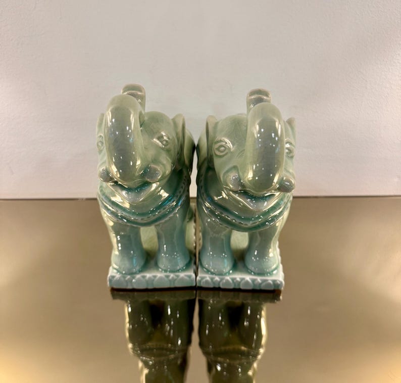 May include: A pair of matching ceramic elephant figurines featuring a glossy, light green celadon glaze. Each elephant is depicted in a standing position with its trunk raised upward, resting on a rectangular base with a decorative scalloped edge.