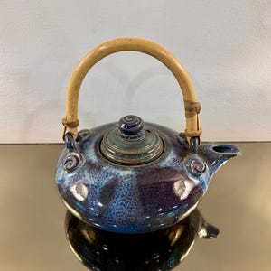 May include: A ceramic teapot with a deep blue and mottled light blue glaze, featuring a rounded body, a matching lid with a small knob, and a curved spout. The teapot has a light-colored, arched bamboo handle attached to two ceramic loops.