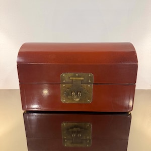 May include: A rectangular, dark red lacquered wooden chest with a curved, domed lid. The front features a central brass latch plate with a decorative, scalloped shape and a horizontal pin mechanism, providing a secure closure for the storage box.