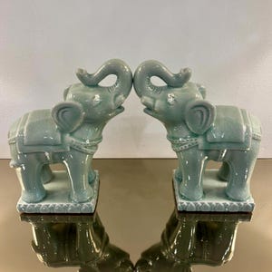 May include: A pair of light green ceramic elephant figurines standing on rectangular bases. Each elephant features a raised trunk, a saddle-like detail on its back, and a visible crackle glaze finish across the entire surface of the ceramic.