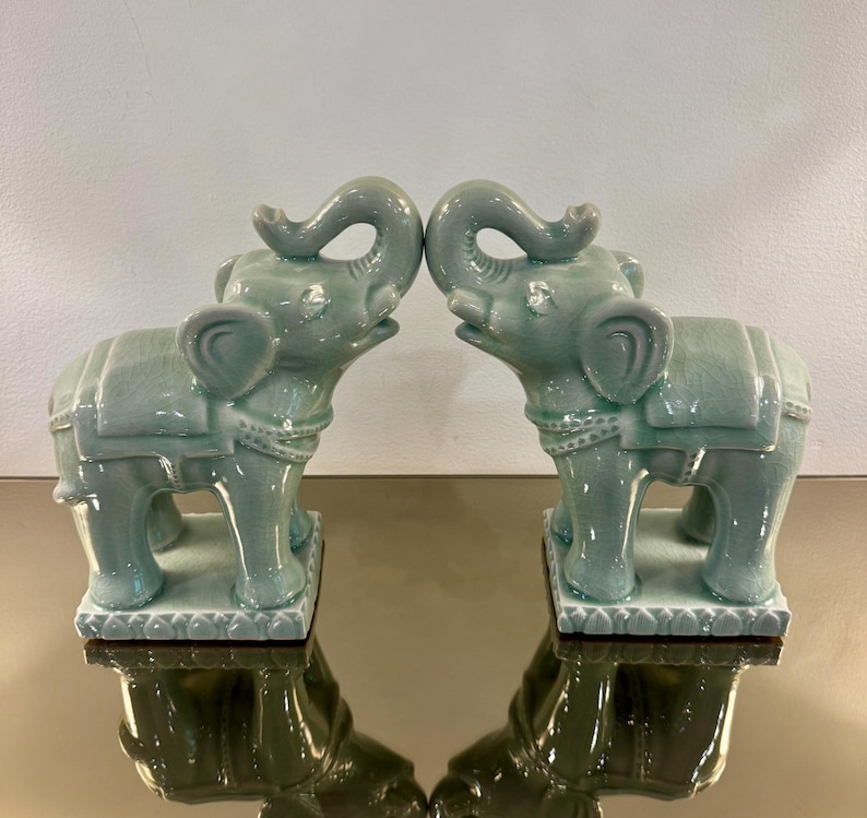 May include: A pair of light green ceramic elephant figurines standing on rectangular bases. Each elephant features a raised trunk, a saddle-like detail on its back, and a visible crackle glaze finish across the entire surface of the ceramic.