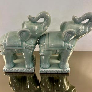 May include: Two matching celadon green ceramic elephant figurines standing on rectangular bases. Each elephant features a glossy crackle glaze finish, a raised trunk, and a decorative saddle blanket with intricate detailing on the back and sides.