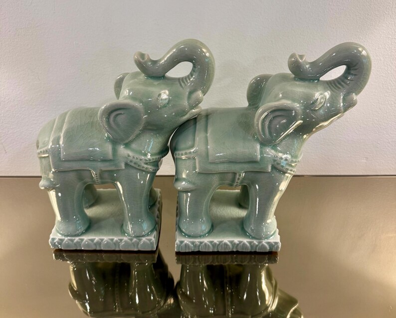 May include: Two matching celadon green ceramic elephant figurines standing on rectangular bases. Each elephant features a glossy crackle glaze finish, a raised trunk, and a decorative saddle blanket with intricate detailing on the back and sides.