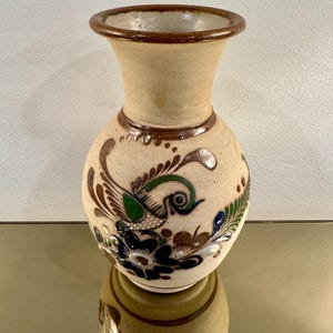 May include: A beige ceramic vase with a round body and a flared neck. The vase is adorned with a hand-painted design featuring a bird, flowers, and foliage in shades of brown, green, blue, and white. The rim and base have a dark brown trim.