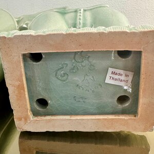May include: A light green ceramic figurine base with a crackle glaze finish. The rectangular underside features four circular holes, an embossed maker mark, and a white rectangular sticker with the printed text Made in Thailand in red ink.