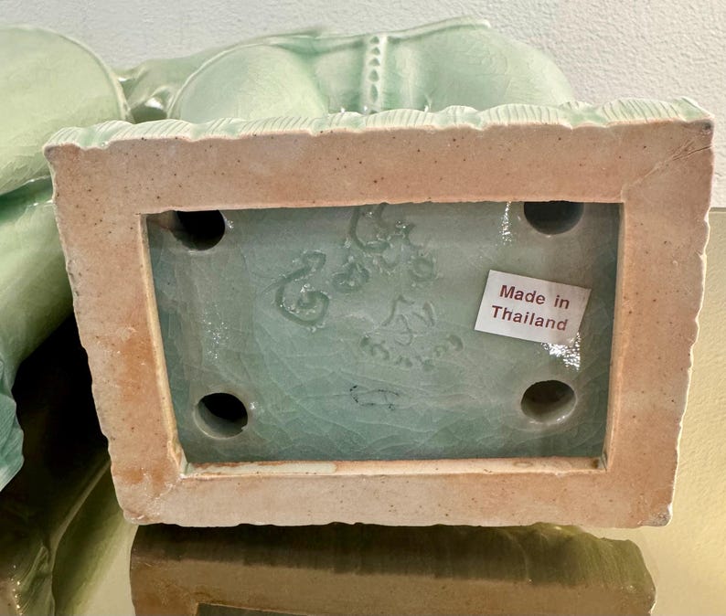 May include: A light green ceramic figurine base with a crackle glaze finish. The rectangular underside features four circular holes, an embossed maker mark, and a white rectangular sticker with the printed text Made in Thailand in red ink.