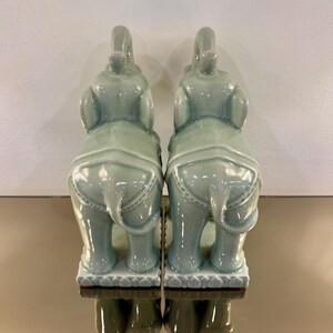 May include: A pair of celadon green ceramic elephant figurines standing on rectangular bases. Each elephant features a raised trunk, large ears, and a decorative saddle blanket pattern on its back. The glossy glaze has a subtle crackle finish.